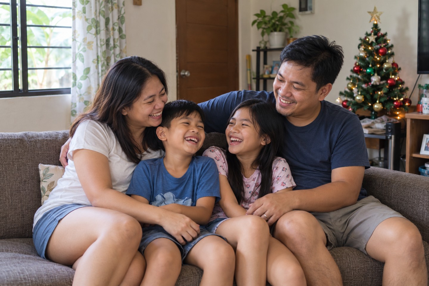 A Filipino family laughing together on their sofa beside a small Christmas tree in a simple home.